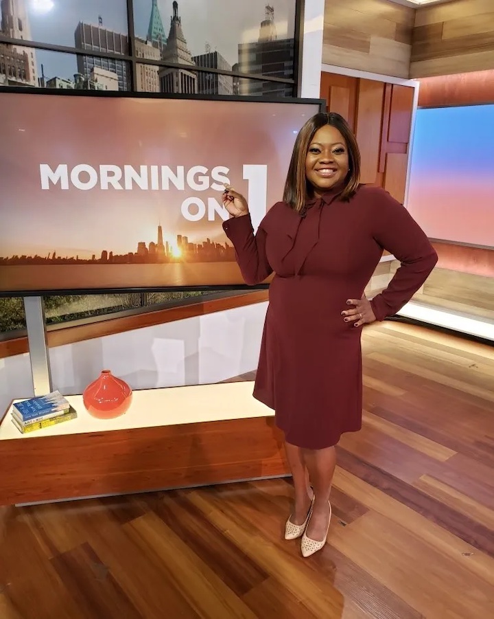 Ruschell Boone standing and pointing to the screen behind her on Mornings on 1 while smiling at the camera.